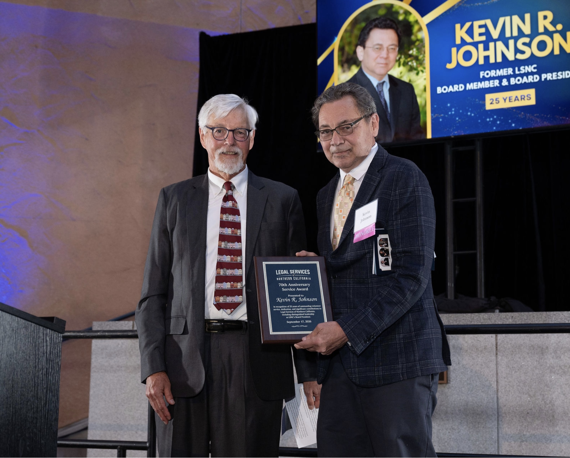 Two men on stage at awards ceremony; one holds a plaque in front of a blue backdrop.