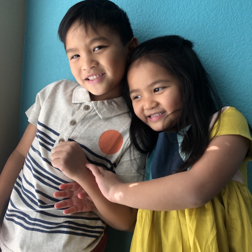 Two children smiling together, one in a striped shirt and the other in a yellow dress, against a blue wall.