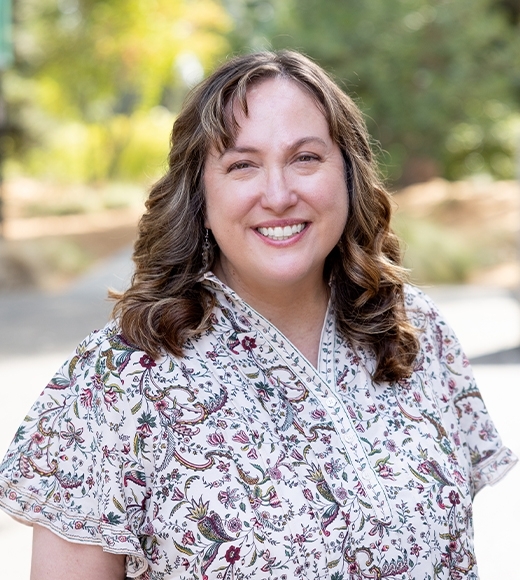 A smiling woman with curly brown hair wearing patterned blouse outdoors.