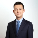 Young man in a business suit with a blue tie, standing against a light background.