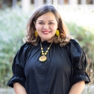 Professor Raquel Aldana wearing a black dress with a gold necklace. She is standing in the King Hall courtyard and smiling at the camera.